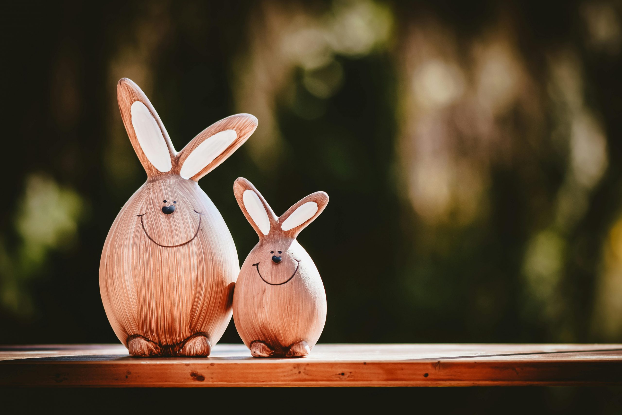 Kein Blasrohrschießen in der Osterwoche Two adorable wooden bunny figurines with happy faces on a table against a blurred outdoor background.